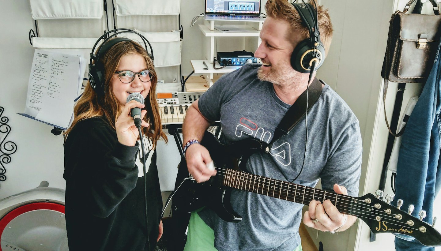 Man speelt elektrische gitaar terwijl meisje zingt in microfoon in thuisstudio.
